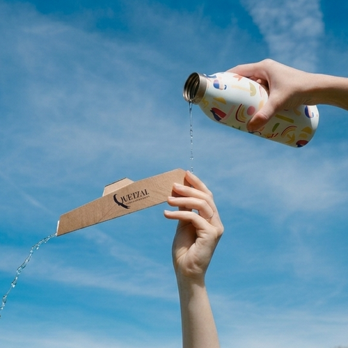 Eau coulant d’une bouteille vers un emballage en carton Mon Quetzal - Compact plié, tenu par deux mains contre un ciel bleu.